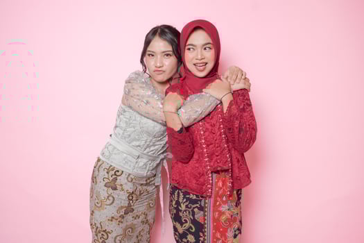 Happy two Asian young woman as close friends wearing modern kebaya and hugging each other showing friendly gesture, isolated over pink background. photo