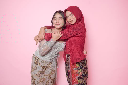 Happy two Asian young woman as close friends wearing modern kebaya and hugging each other showing friendly gesture, isolated over pink background. photo