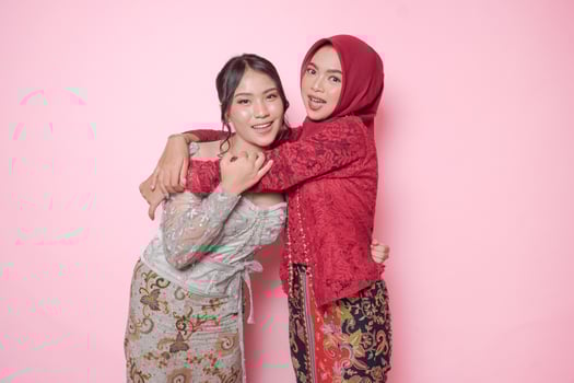 Happy two Asian young woman as close friends wearing modern kebaya and hugging each other showing friendly gesture, isolated over pink background. photo