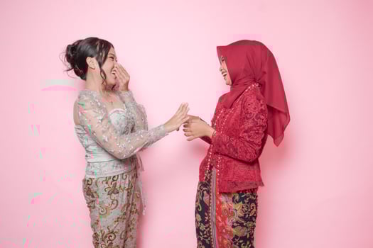 Surprised Asian women wearing modern kebaya close friends having shocking conversation, isolated over pink background. photo