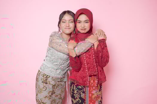 Happy two Asian young woman as close friends wearing modern kebaya and hugging each other showing friendly gesture, isolated over pink background. photo