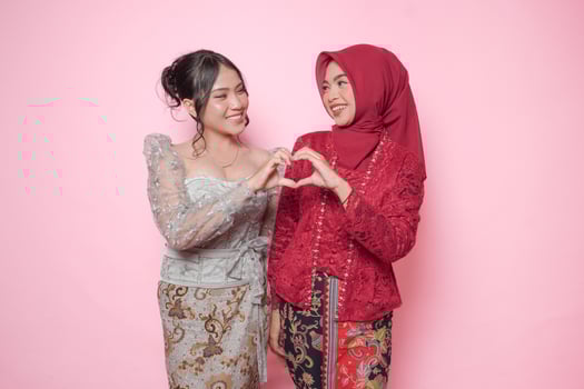 Excited two Asian young woman wearing modern kebaya making heart shape gesture together, isolated over pink background. photo