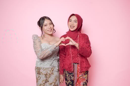 Excited two Asian young woman wearing modern kebaya making heart shape gesture together, isolated over pink background. photo