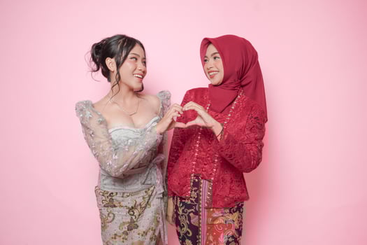 Excited two Asian young woman wearing modern kebaya making heart shape gesture together, isolated over pink background. photo