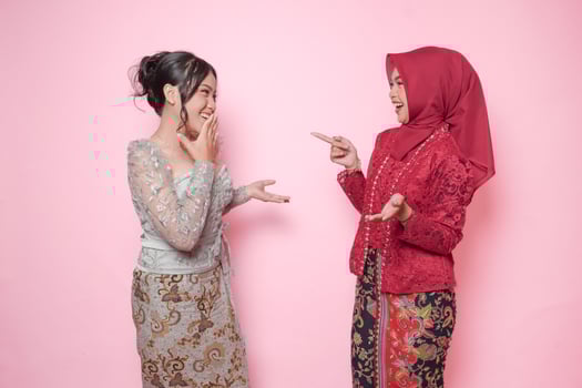 Two Asian women as close friends wearing modern kebaya having a fun conversation, isolated over pink background. photo