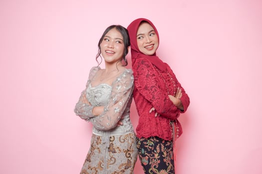 Two smiling Asian women wearing modern kebaya dress posing and leaning on each other back, isolated by pink background. photo