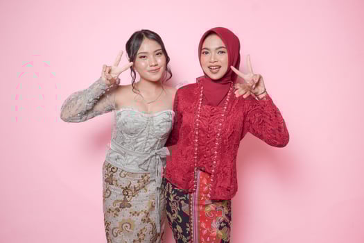 Two joyful Asian young woman wearing modern kebaya showing peace sign, isolated over pink background. photo