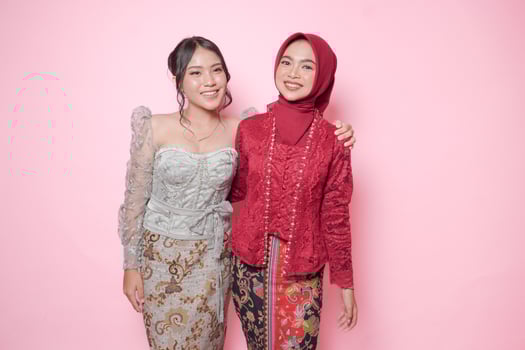 Two cheerful Asian young woman wearing modern kebaya hugging each other showing friendly gesture, isolated over pink background. photo