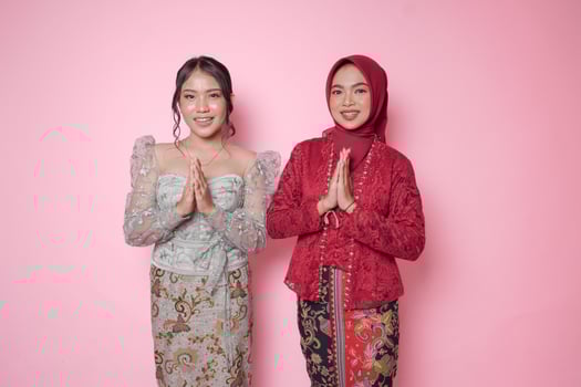 Two beautiful Asian women wearing modern kebaya dress doing welcome greetings hand gesture over isolated pink background. photo