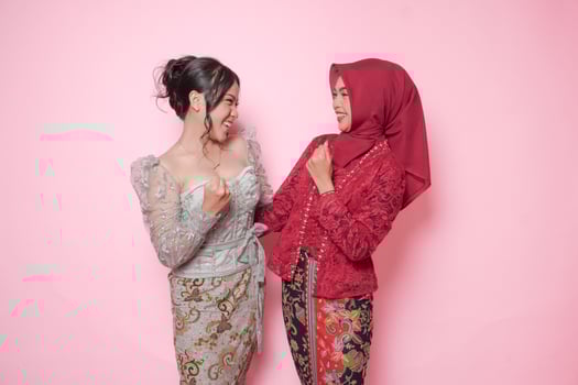 Two excited Asian women wearing modern kebaya dress with a happy successful expression looking at each other, isolated by pink background. photo