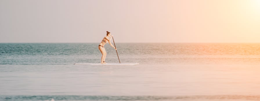 Sea woman sup. Silhouette of happy middle aged tanned woman in rainbow bikini, surfing on SUP board, confident paddling through water surface. Idyllic sunset. Active lifestyle at sea or river. photo