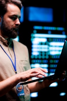 Serviceman doing system analysis, typing data on laptop of needed adjustments in supercomputers server farm housing advanced data storage infrastructure supporting critical IT workloads, close up photo