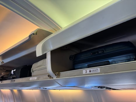 Overhead storage compartment inside airplane cabin filled with various carry on luggage, including black and white suitcases, backpacks, bags, with compartment doors open, visible seat row numbers. photo