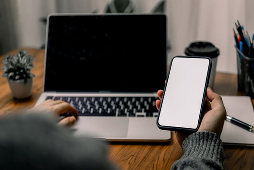 Person using a smartphone while sitting at a desk with a laptop, suggesting multitasking or mobile communication. The screen of the smartphone is blank. photo