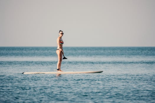 Sea woman sup. Silhouette of happy middle aged tanned woman in rainbow bikini, surfing on SUP board, confident paddling through water surface. Idyllic sunset. Active lifestyle at sea or river. photo