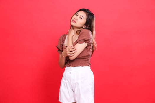 Asian woman's expression of shoulder pain looking at the camera wearing a brown blouse top and shorts on a red background. for health, lifestyle and advertising concepts photo