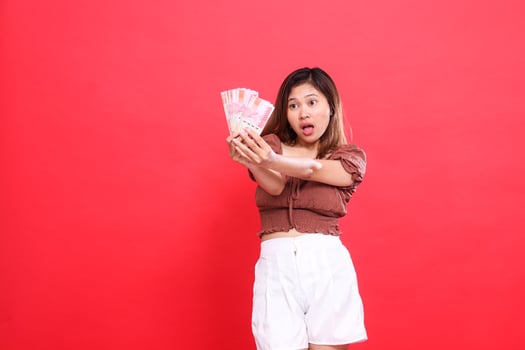 gesture of a young indonesia woman shocked candid with both hands holding rupiah money in front of her wearing a brown blouse top with a red background. for transaction, technology and advertising photo