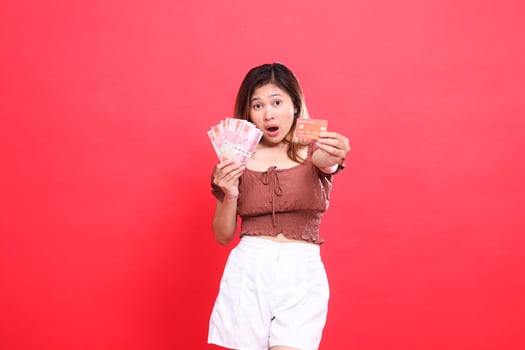 Shocked Asian woman holding Rupiah currency and debit credit card in front of her wearing a brown blouse with a red background. for transaction, technology and advertising concepts photo