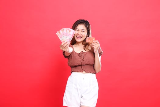 Happy indonesia woman holding rupiah money in front of her and a debit credit card wearing a brown blouse on a red background. for transaction, technology and advertising concepts photo