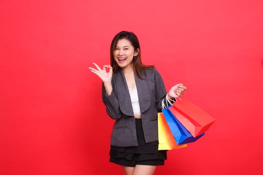 gesture of a cheerful indonesia businesswoman carrying a shopping paper bag, hand a sign of okay good, wearing a formal jacket on a red background. for commercial, lifestyle and sales concepts photo