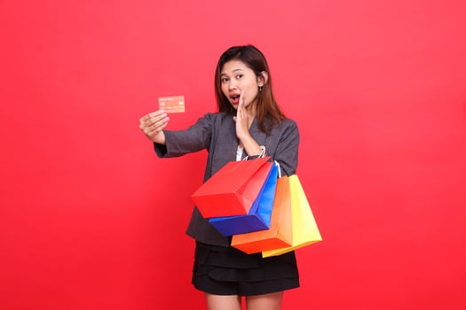 Asian office woman shocked to the left carrying a shopping paper bag while holding a credit debit card to the front wearing a formal jacket with a red background. for transaction and business photo