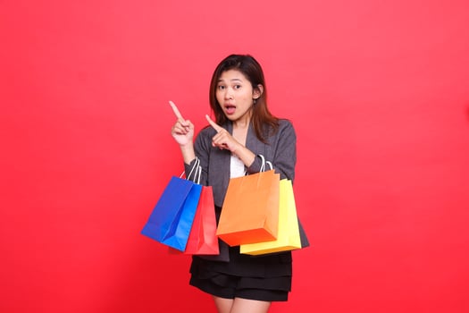 Asian office girl expression is shocked, carrying a shopping paper bag, hand pointing up right, wearing a formal jacket on a red background. for commercial, lifestyle and sales concepts photo