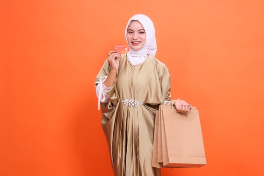 Cheerful young indonesia woman carrying a brown paper bag and holding a member card, debit credit card wearing a kaftan Muslim dress with a hijab. Fashion concept, transaction, promotion photo