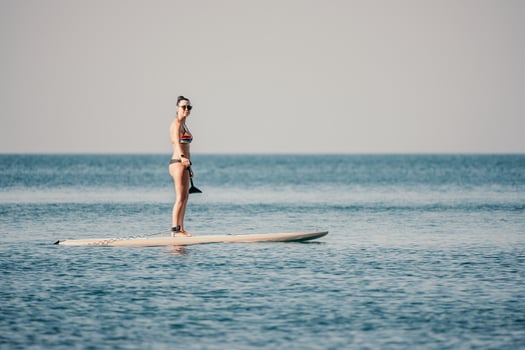 Sea woman sup. Silhouette of happy middle aged tanned woman in rainbow bikini, surfing on SUP board, confident paddling through water surface. Idyllic sunset. Active lifestyle at sea or river. photo