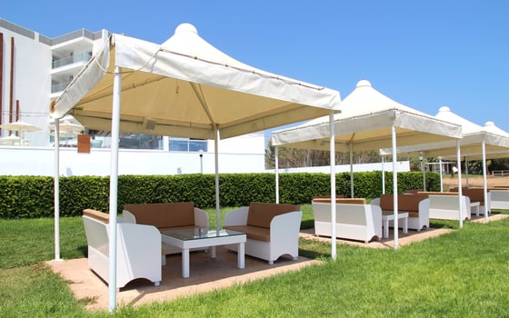 Outdoor lounge area with white tents and modern furniture in a sunny resort setting, featuring green grass and a hedge, against a clear blue sky and a contemporary building in the background photo