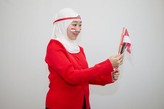 Woman in white hijab wearing red sweater. celebrating Indonesia's independence day. holding a flag and taking a selfie with a cellphone. isolation on white background. photo