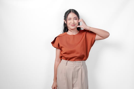 Cute young Asian woman in a brown shirt making a heart gesture on her cheek, isolated over white background. photo