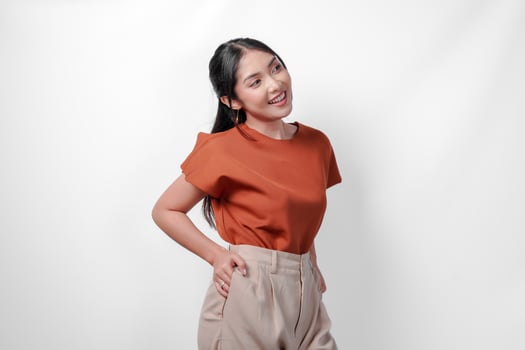 Beautiful young Asian woman in brown shirt and taupe pants placing hands on hip with confident expression, isolated by white background. photo