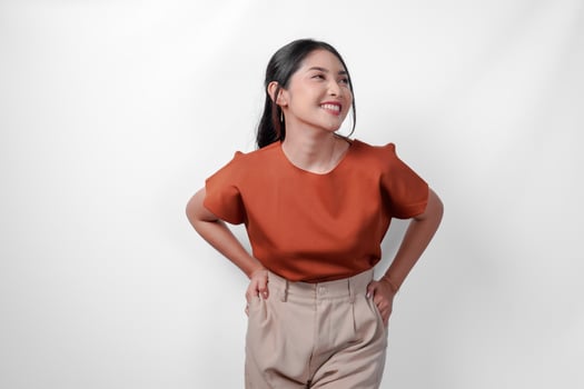 Beautiful young Asian woman in brown shirt and taupe pants placing hands on hip with confident expression, isolated by white background. photo