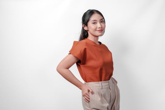 Beautiful young Asian woman in brown shirt and taupe pants placing hands on hip with confident expression, isolated by white background. photo