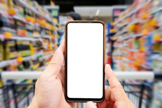 A person's hand holds a mobile phone, with a shopping basket on the screen. In the background, a pretty woman selects digital products, in a blur. Mock up. photo