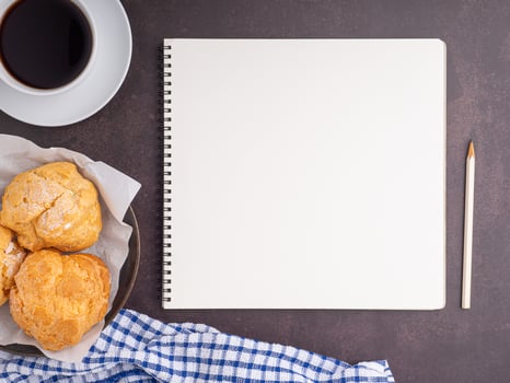The top view of a notepad with a pencil, choux cream on a plate, and a white coffee cup are placed on dark brown background. Space for text. Concept of writing and relaxation photo