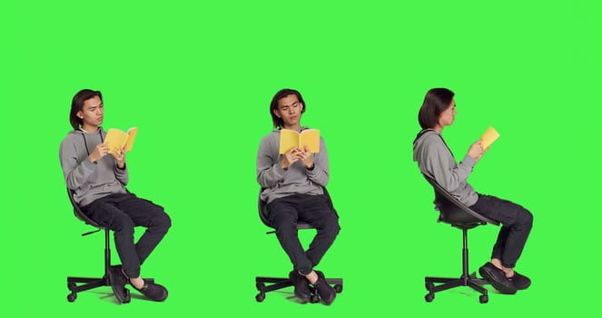 Asian guy reading literature book over greenscreen backdrop, enjoying lecture while he sits on chair in studio. Person reads fiction novel story, being interested in classic leisure activities. photo