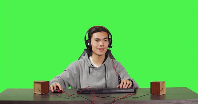 POV of asian guy playing at pc desktop, sitting against full body greenscreen backdrop. Young gamer having fun with players online in gaming competition, wearing headphones at workstation. photo