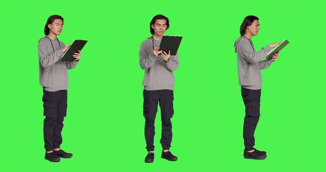 Asian adult reading information on files, thinking about annotations on clipboard papers or taking notes for finalization. Young guy writing on documents, standing against greenscreen. photo