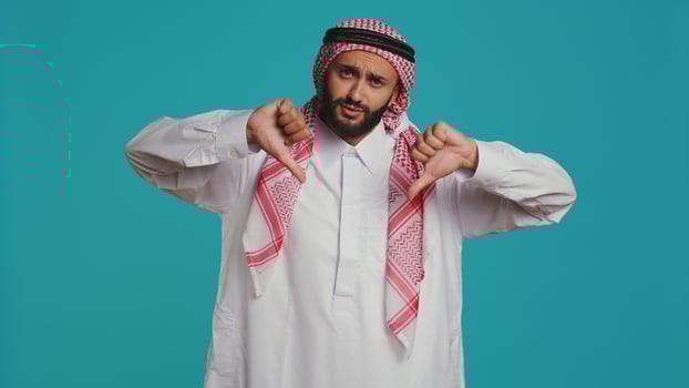 In front of background, muslim adult gives thumbs up indicating fulfillment and happiness while dressed in traditional arabic clothes. Young man wearing religious islamic clothing, like sign. photo