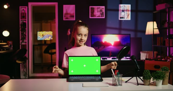 Cheerful child in home studio presenting advertisement from sponsoring brand on green screen laptop. Gen Z girl using chroma key device to present promotion from media partner to followers photo