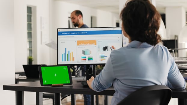 Analyst working at office desk with greenscreen shown on tablet, looking at blank copyspace layout on device while she checks sales data. Employee uses chromakey template. Handheld shot. photo
