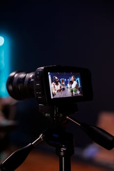 Detailed view of professional camera recording male journalist with notebook interviewing famous blogger. Selective focus of digital device on tripod filming black couple doing live talk show. photo