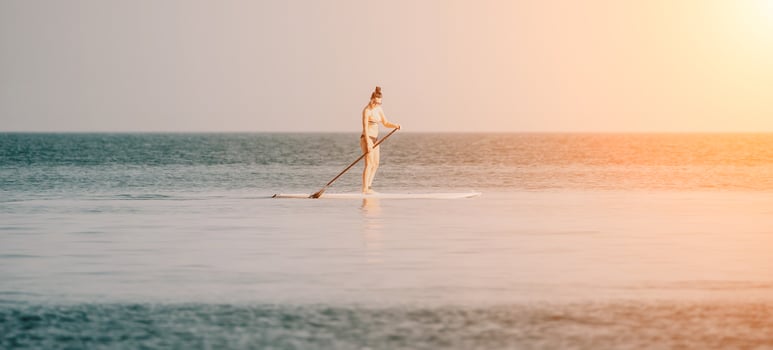 Sea woman sup. Silhouette of happy middle aged woman in rainbow bikini, surfing on SUP board, confident paddling through water surface. Idyllic sunset. Active lifestyle at sea or river. photo