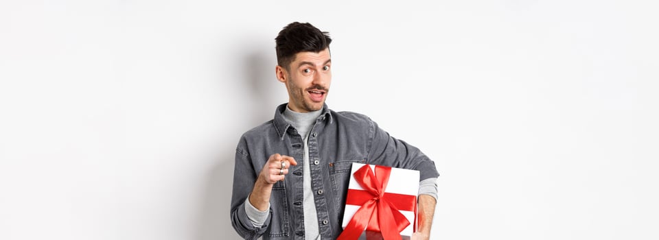 Portrait of romantic guy buying gift for Valentines day and pointing at you, inviting to special deal shopping, standing with present on white background photo