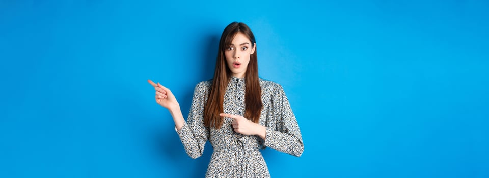 Surprised natural girl in dress say wow, pointing fingers right at promo and look impressed, asking question about advertisement, blue background photo