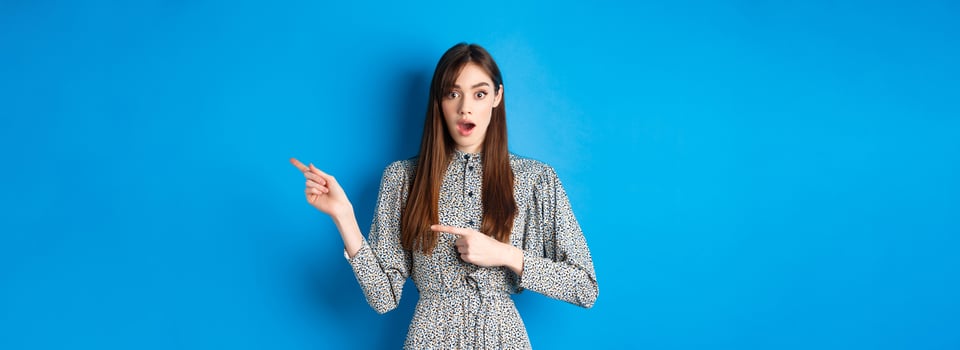 Wow look that way. Excited young woman with long hair and trendy dress, drop jaw and pointing right at logo, showing awesome news, blue background photo