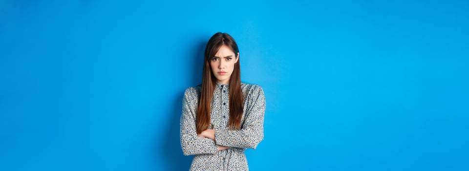 Angry frowning woman in dress, cross arms on chest and feel offended about something unfair, standing upset against blue background photo