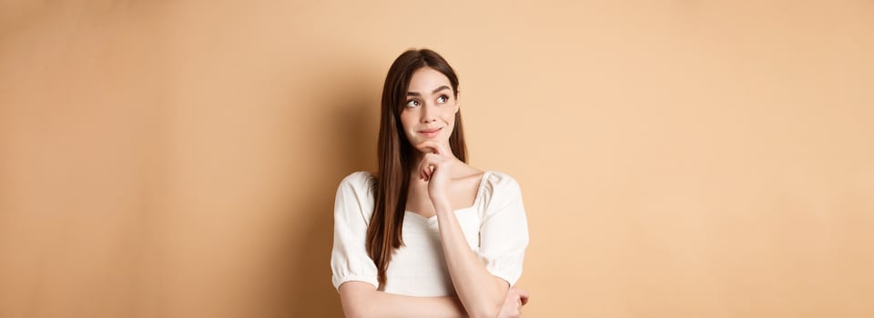 Very interesting. Pensive cute girl thinking and looking at upper left corner logo, smiling pleased, having good idea, standing on beige background photo