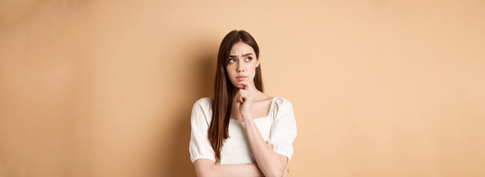 Worried young woman staring pensive aside, touching chin and thinking, pondering plan, looking for ideas, standing on beige background photo